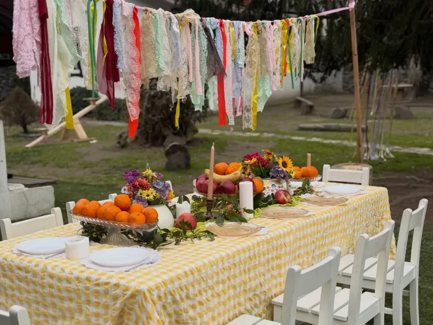 Bahçede uzun, sarı pötikareli sofra: meyveler, çiçekler, mumlar ve beyaz çocuk sandalyeleri
