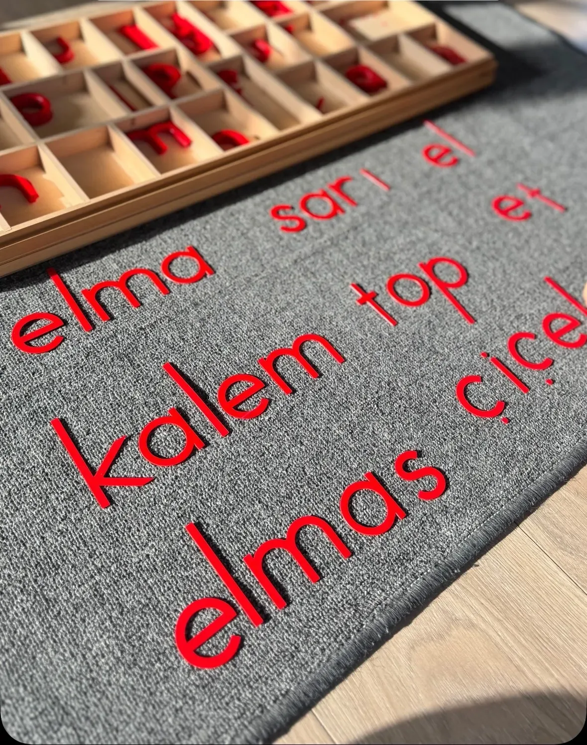 Kırmızı harfli hareketli alfabe kutusu; gri keçe üzerinde dizilmiş kelimeler: elma, sarı, el, et, kalem, top, elmas, çiçek
